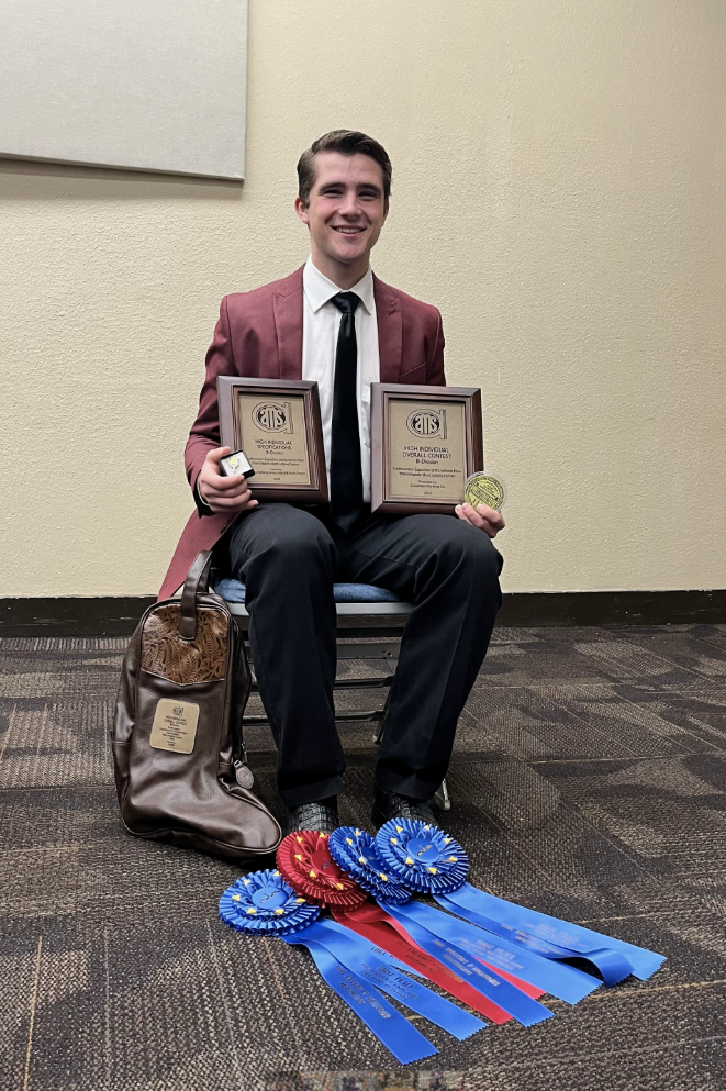 Zach Holescher sitting in a chair holding two awards with award ribbons also laid at his feet on the floor.